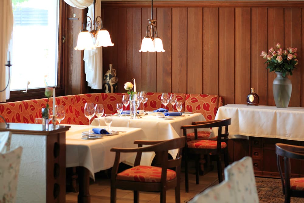 Gemütlicher Restaurantbereich mit Holzvertäfelung, gedeckten Tischen mit weißen Tischdecken, roten Stühlen und Sofas sowie Blumendekoration.