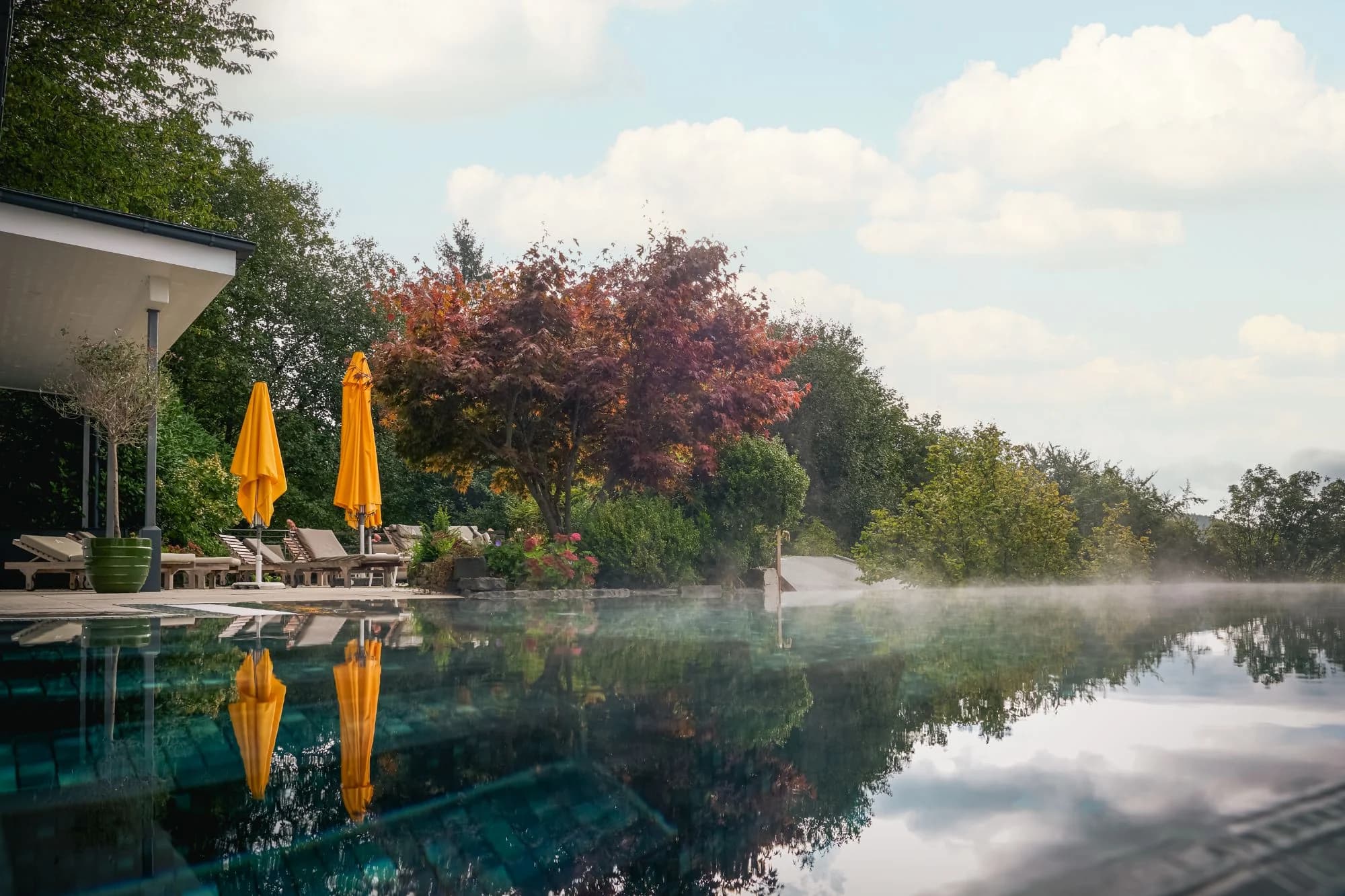 Dampfender Infinity-Pool im Herbst, gelbe Sonnenschirme und rotes Laub spiegeln sich im Wasser